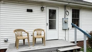 Une maison avec un porche à l’avant, deux chaises en plastique, une porte et un coffret de raccordement électrique.