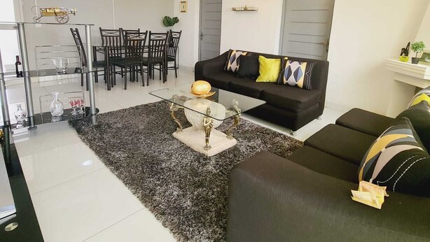 Een moderne woonkamer met een zwarte bank, een glazen salontafel en een kroonluchter. Er is een eethoek met een tafel en stoelen.