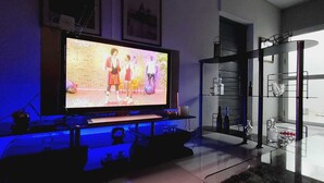 Een woonkamer met een grote flatscreen-tv, een glazen salontafel en een moderne keuken met een koelkast en uitgestalde wijnglazen.