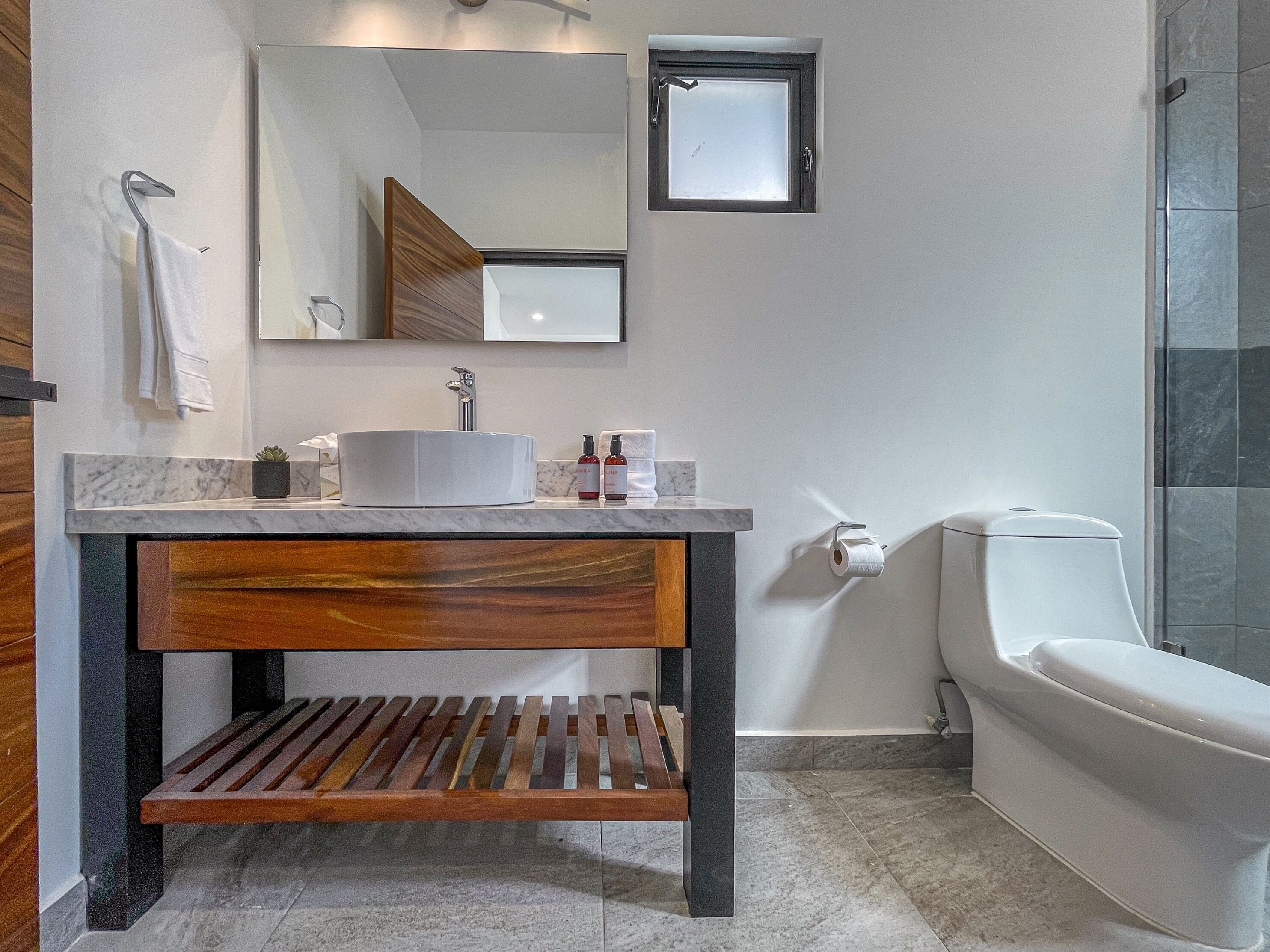 Une salle de bain moderne avec un plan de toilette en marbre, un banc en bois, un lavabo blanc et une petite fenêtre au-dessus des toilettes.