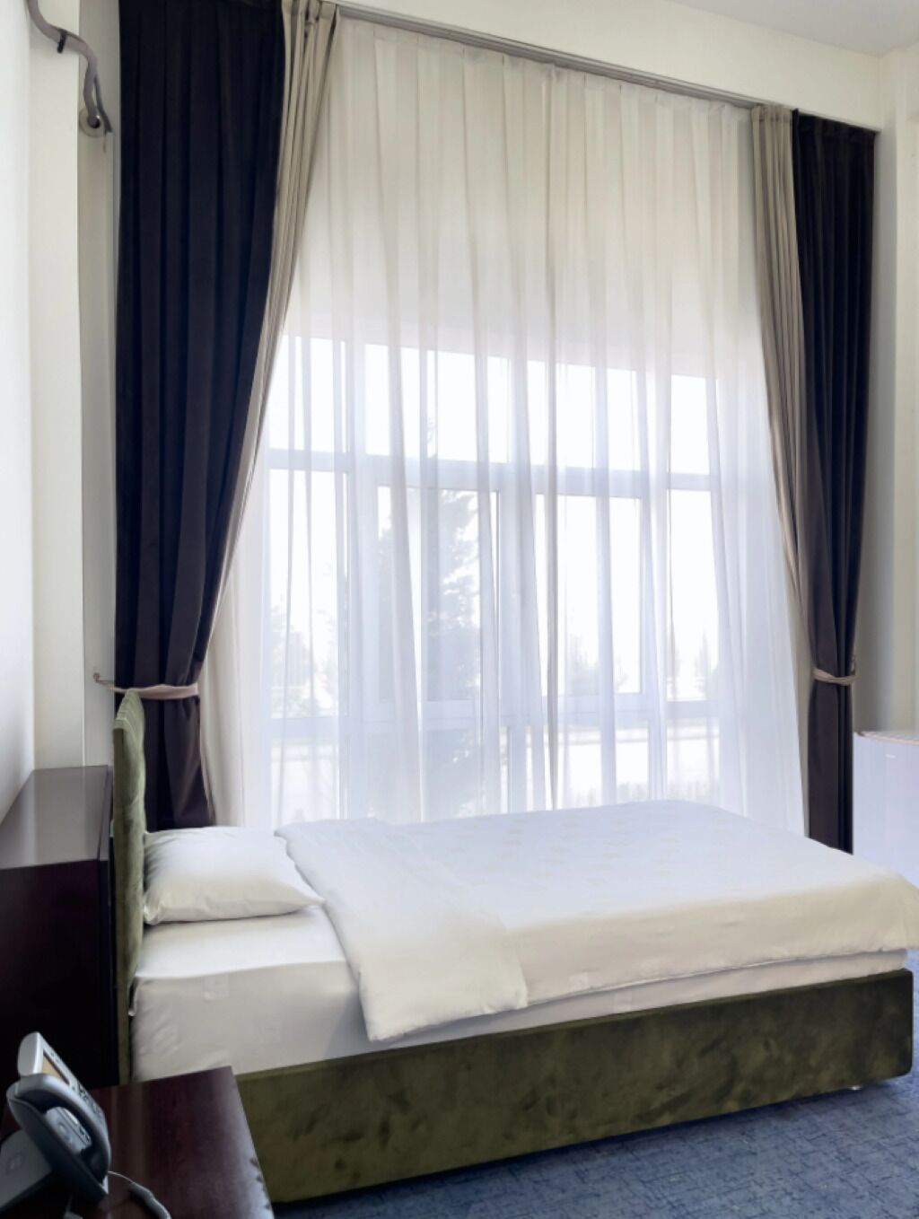 Une chambre d’hôtel avec un lit, un bureau et une fenêtre ornée de rideaux légers.