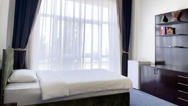 A bedroom with a bed, a bedside table, a wardrobe, and a window with sheer curtains. - VIP Room 2 - Ussat Hotel (Ashgabat)
