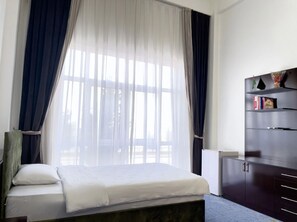 Une chambre à coucher comprenant un lit, une table de chevet, une armoire et une fenêtre avec des rideaux légers.
