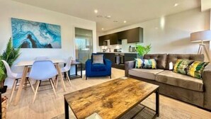 Een moderne woonkamer met een houten salontafel, een blauwe bank en een eethoek met witte stoelen.