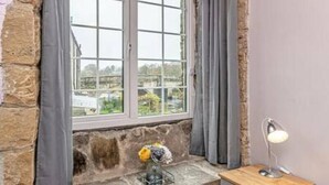A room with a stone wall, a window with curtains, a small table with a lamp, and a vase with flowers.