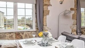 A dining area with a table set for four, a flower vase, and a view of a stone wall and a bridge outside the window.