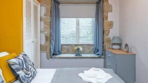 A bedroom with a stone wall, a window with curtains, a bed with a gray bedspread, a bedside table with a lamp, and a yellow pillow.