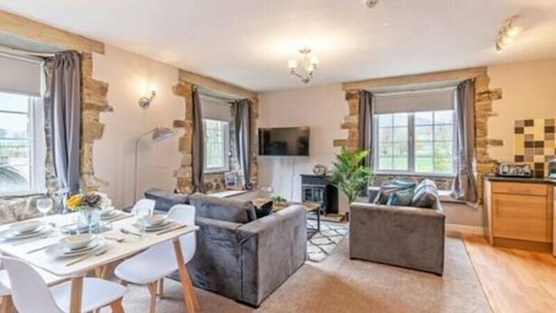 A modern living room with a dining area, grey sofas, a flat-screen TV, and a kitchen with wooden cabinets.