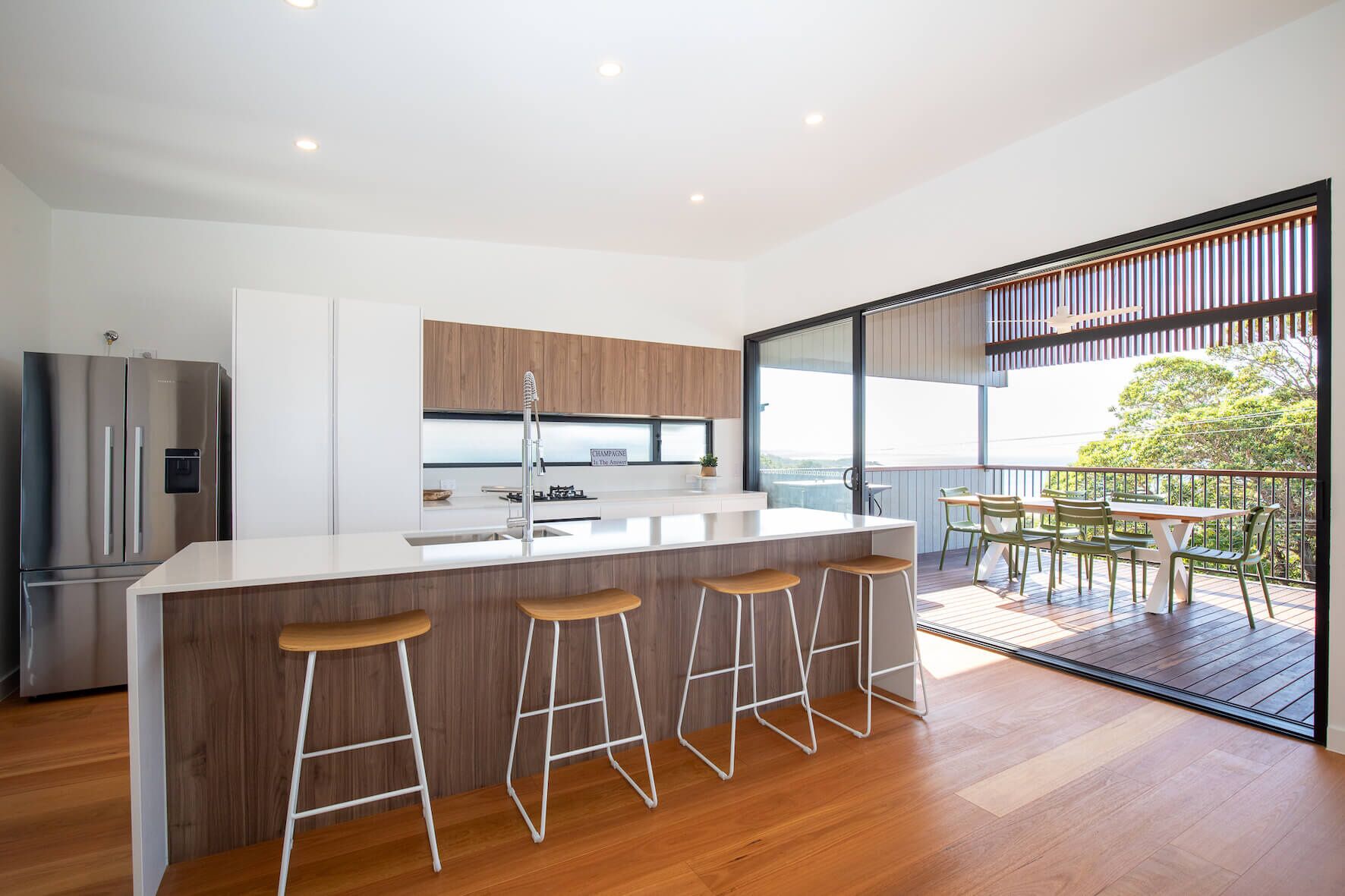 A modern kitchen with a central island, bar stools, and a view of a balcony with outdoor seating.