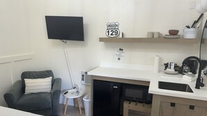 Een compacte kitchenette met een magnetron, gootsteen en kleine koelkast. Aan de muur hangt een flatscreen-tv en boven de aanrecht is een bordje.