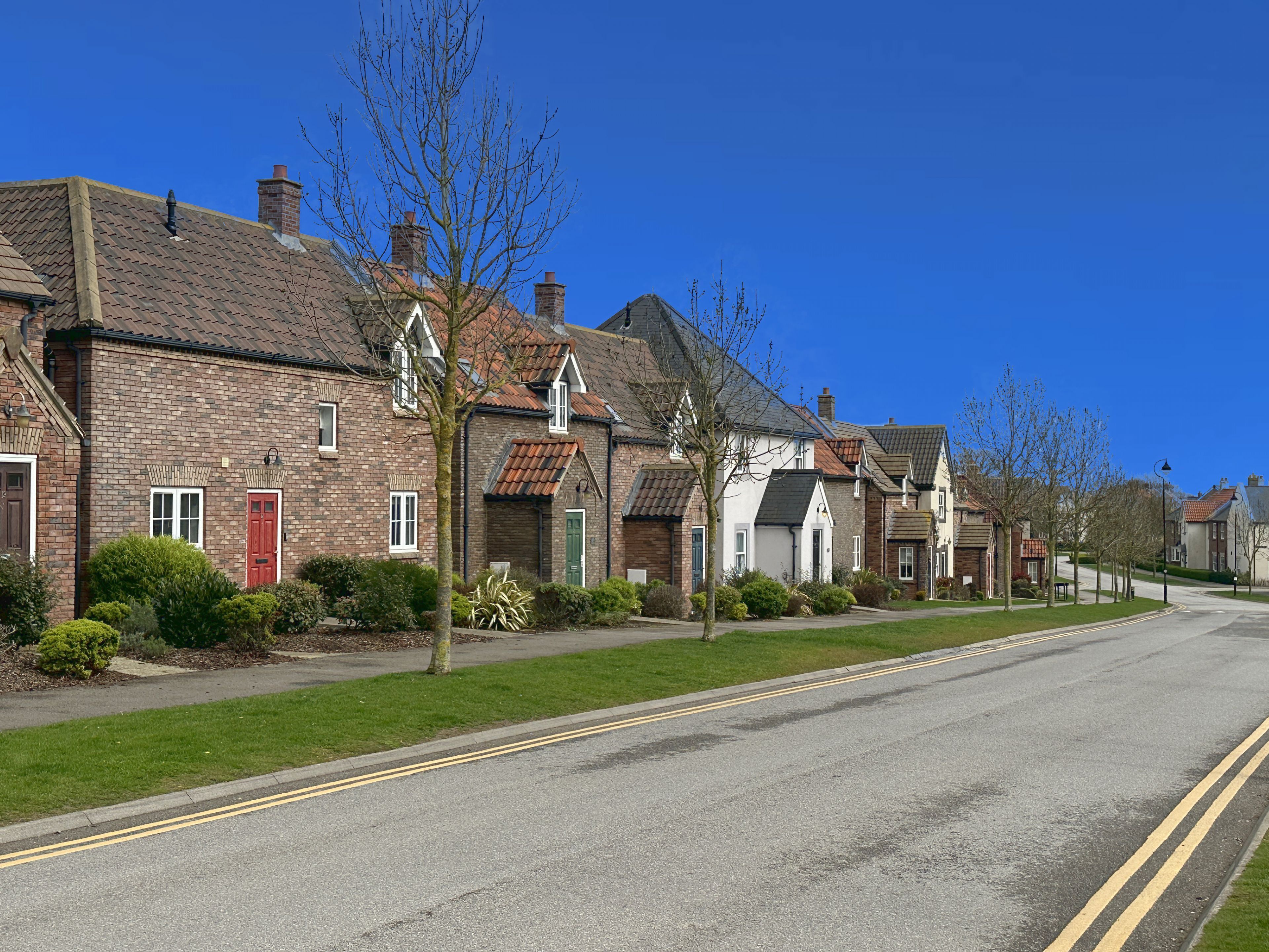 Une rangée de maisons traditionnelles en briques, avec des toits à deux versants et des cheminées, un ciel bleu dégagé et une route à deux voies délimitée par des lignes jaunes continues.
