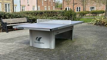 A ping pong table with the brand 'Cornilleau' in a public outdoor area with a paved surface, a bench, and buildings in the background.