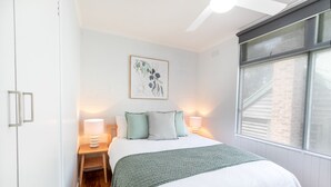 Un dormitorio ordenado con cama, lámparas de noche, una obra de arte en la pared y una ventana con persianas.