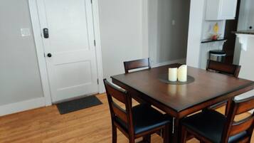 Un comedor con una mesa de madera y cuatro sillas, una puerta blanca y una zona de cocina al fondo.