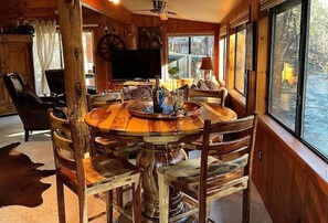A wooden interior with a round dining table surrounded by chairs, a television, and a large window offering a view of the outdoors.