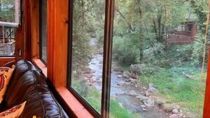 A cozy cabin interior with a large window overlooking a forested area and a stream. - Waterfront luxurious midcentury Log cabin (Payson)