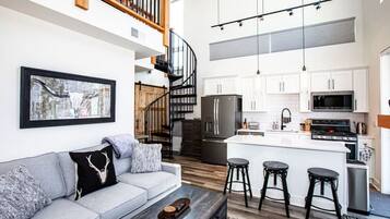 A modern kitchen with white cabinets, a black refrigerator, and a stainless steel stove. There is a gray sofa with a black throw pillow and a wooden coffee table. A staircase with wooden railings leads to an upper level.