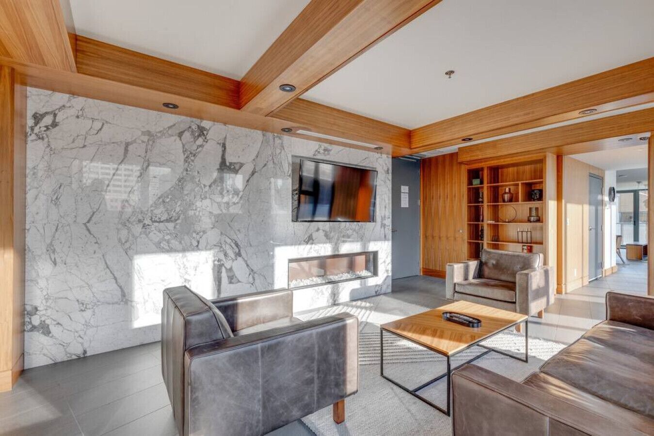 A modern living room with a marble wall, wooden ceiling beams, a flat-screen TV, and a built-in bookshelf.
