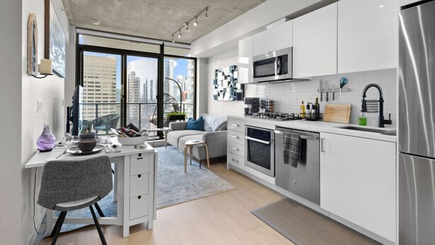 A modern kitchen with white cabinets, stainless steel appliances, and a dining area with a gray chair and table. - Cozy DT Fantastic Amenities View Private Parking (Calgary)