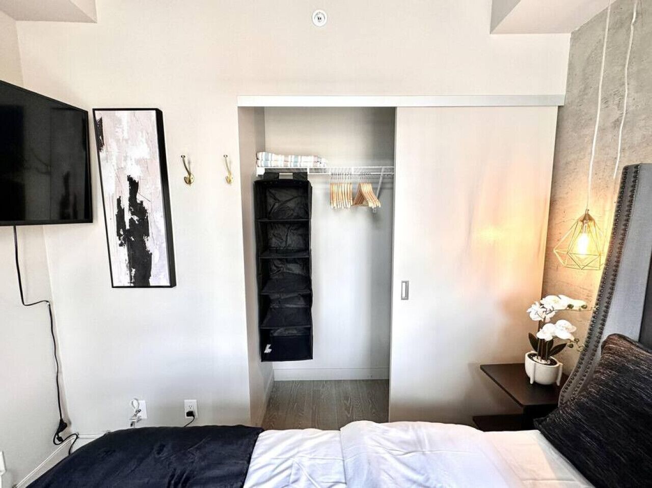 Une chambre moderne avec un lit, un téléviseur fixé au mur, une armoire avec des vêtements suspendus et une table de chevet avec une lampe et une plante.