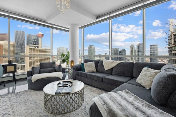 A modern living room with a large window, a grey sofa, a coffee table, and a view of the city skyline. - Urbanest - Spectacular Views & Amenities Downtown (Calgary)