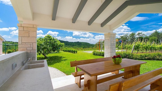 Eine Terrasse im Freien mit Holztisch und Bänken, mit Blick auf einen gepflegten Garten und einen Weinberg.
