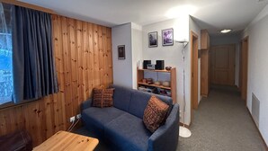 A compact living space with a wooden panel wall, a blue sofa with patterned cushions, a wooden coffee table, and a bookshelf with decorative items. - Crêtes X2 232 - SUNNY apartment 6 pers (Veysonnaz)