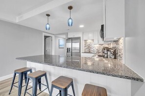 A modern kitchen with granite countertops, stainless steel appliances, and a backsplash of small tiles. There are three wooden stools at the island. - 3 BR Coastal Getaway w/Pool (Myrtle Beach)