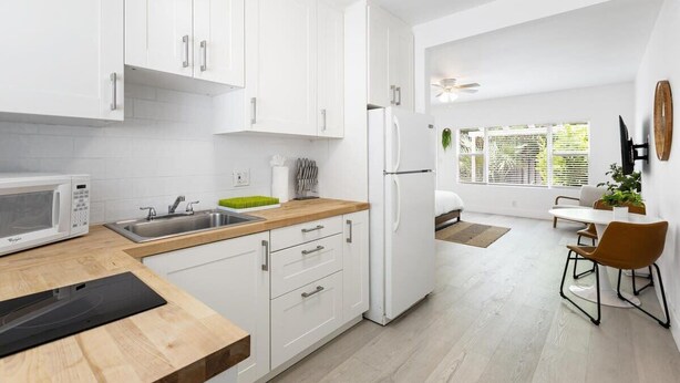 A modern kitchen with white cabinets, a microwave, a sink, and a refrigerator. There is a dining area with a table and chairs.