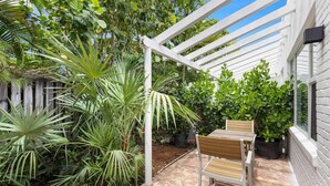 A covered patio with a wooden table and chairs, surrounded by lush greenery and a white pergola.