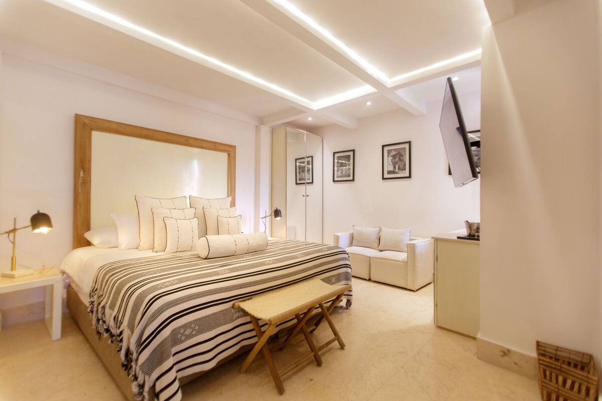 A modern bedroom with a large bed, bedside tables, a bench, a wardrobe, and a flat-screen TV mounted on the wall.