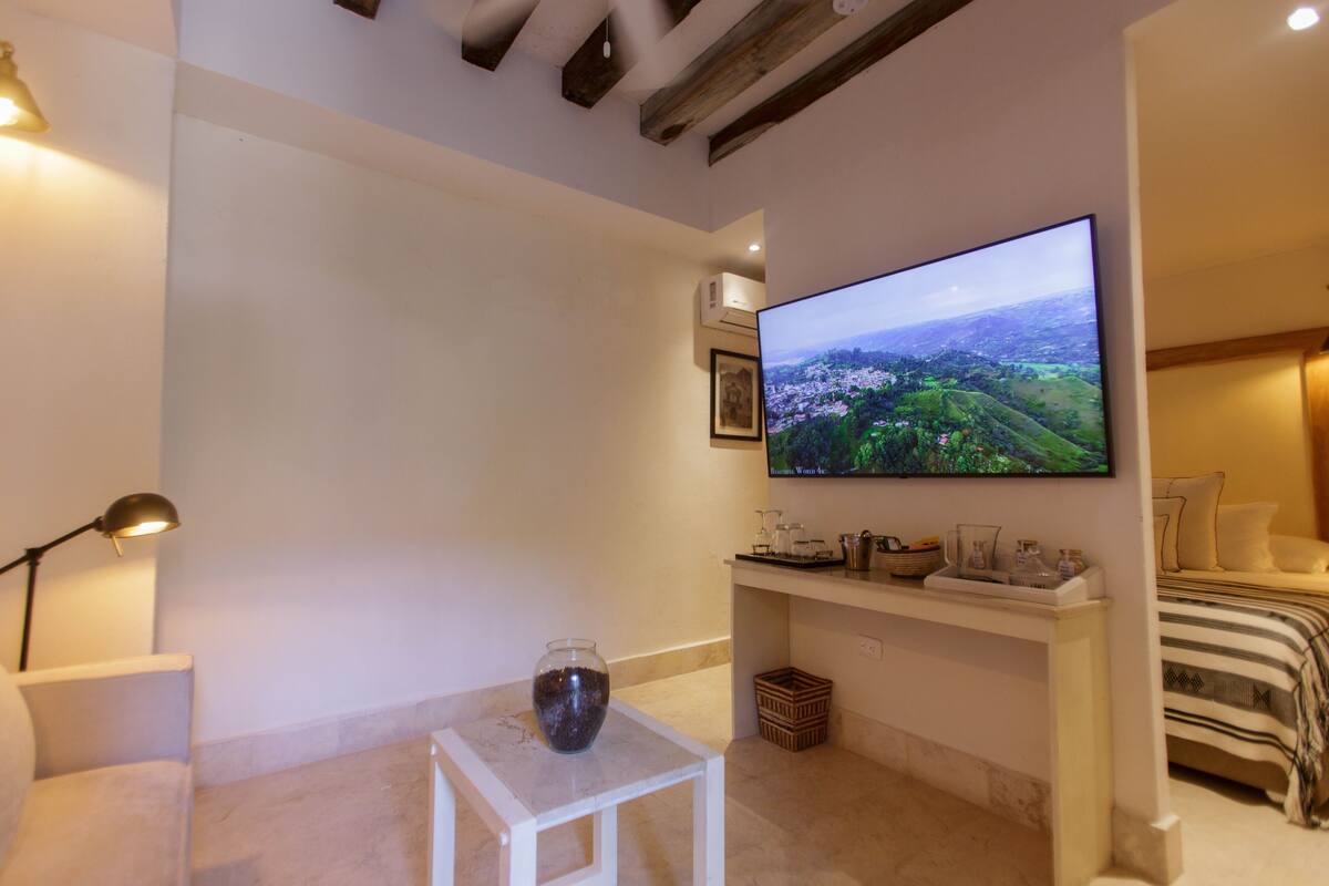 A modern living room with a flat-screen TV mounted on the wall, a small white table with a vase, a lamp, and a bed with a striped blanket.