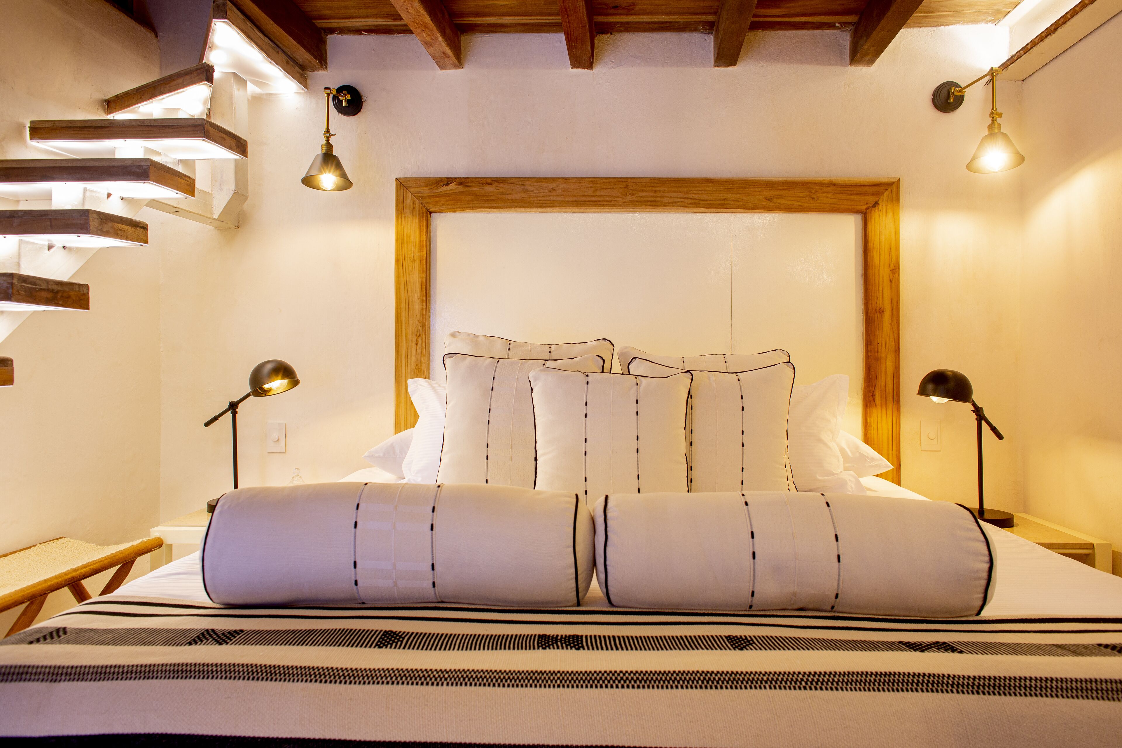 A neatly made bed with a headboard, two bedside lamps, and a wooden staircase with built-in lighting.