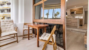 A modern living room with a wooden dining table, two chairs, and a sofa. A large window offers a view of palm trees. - Home2Book Design Apartment Maspalomas Paraíso (Maspalomas)