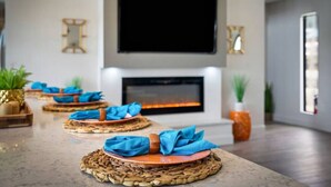 Un salon moderne avec une cheminée, un téléviseur à écran plat fixé au-dessus, et une longue table à manger agrémentée de sets de table en fibres naturelles et de serviettes bleues.