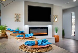 A modern living room with a fireplace, a flat-screen TV mounted above it, and a long dining table set with woven placemats and blue napkins.