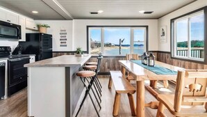 A modern kitchen with a large island, bar stools, and a dining table with benches. There are large windows offering a view of the outdoors.