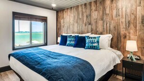 A bedroom with a large bed, a view of the water through a window, and a wooden wall.