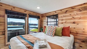 A wooden cabin bedroom with a large bed, multiple pillows, and a view of a lake.