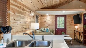 A wooden cabin interior with a kitchen area featuring a sink and countertop, a living room with a sofa and a television, and a door with a window.