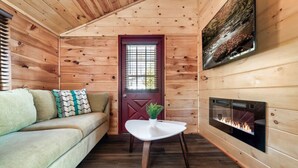 A cozy cabin interior with a wooden interior, a sofa with patterned cushions, a small table with a plant, a fireplace, and a door with a window.