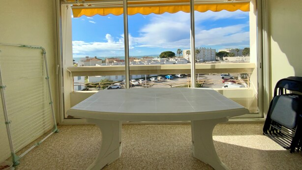 A room with a large window, a white table, and a view of a parking area and buildings. - TELLINES - Appartement  avec petite vue mer (Le Grau-du-Roi)