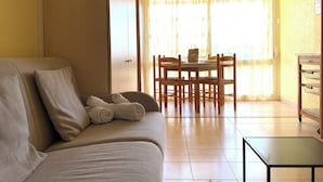 Una sala de estar con un sofá, una mesa de comedor con sillas, una mesita y un estante con objetos.