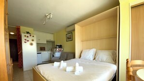 Habitación de hotel con cama, cabecero de madera, silla de madera, kitchenette pequeña y baño.
