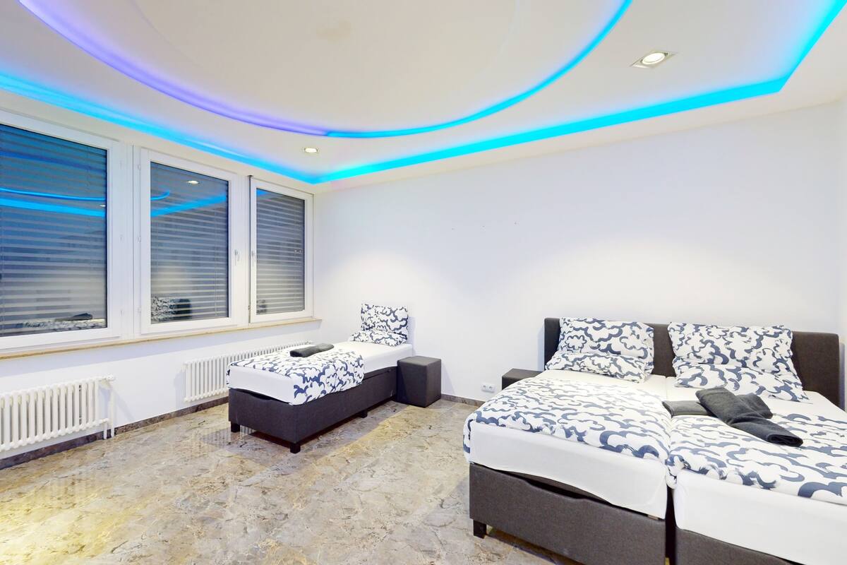 Une chambre moderne avec un grand lit, un lit plus petit et un coin salon. De grandes fenêtres sont équipées de stores, il y a un radiateur et le plafond est doté d’un éclairage bleu.