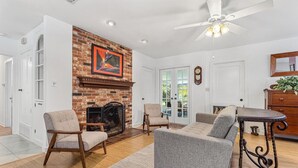 Una sala de estar con chimenea de ladrillo, ventilador de techo y comedor con mesa y sillas.