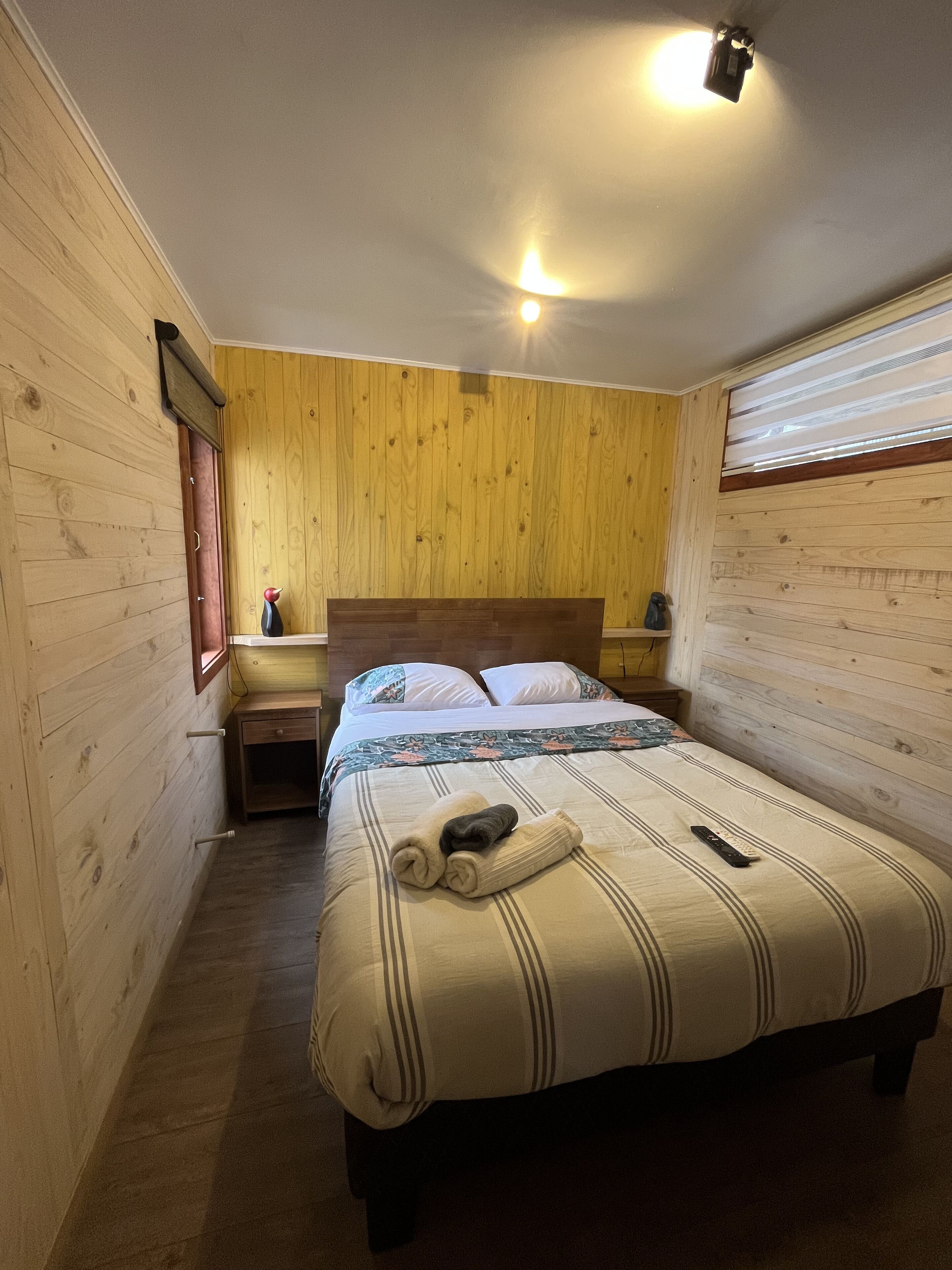 A bedroom with a wooden headboard, a bed with striped bedding, a bedside table with a lamp, and a window with blinds.