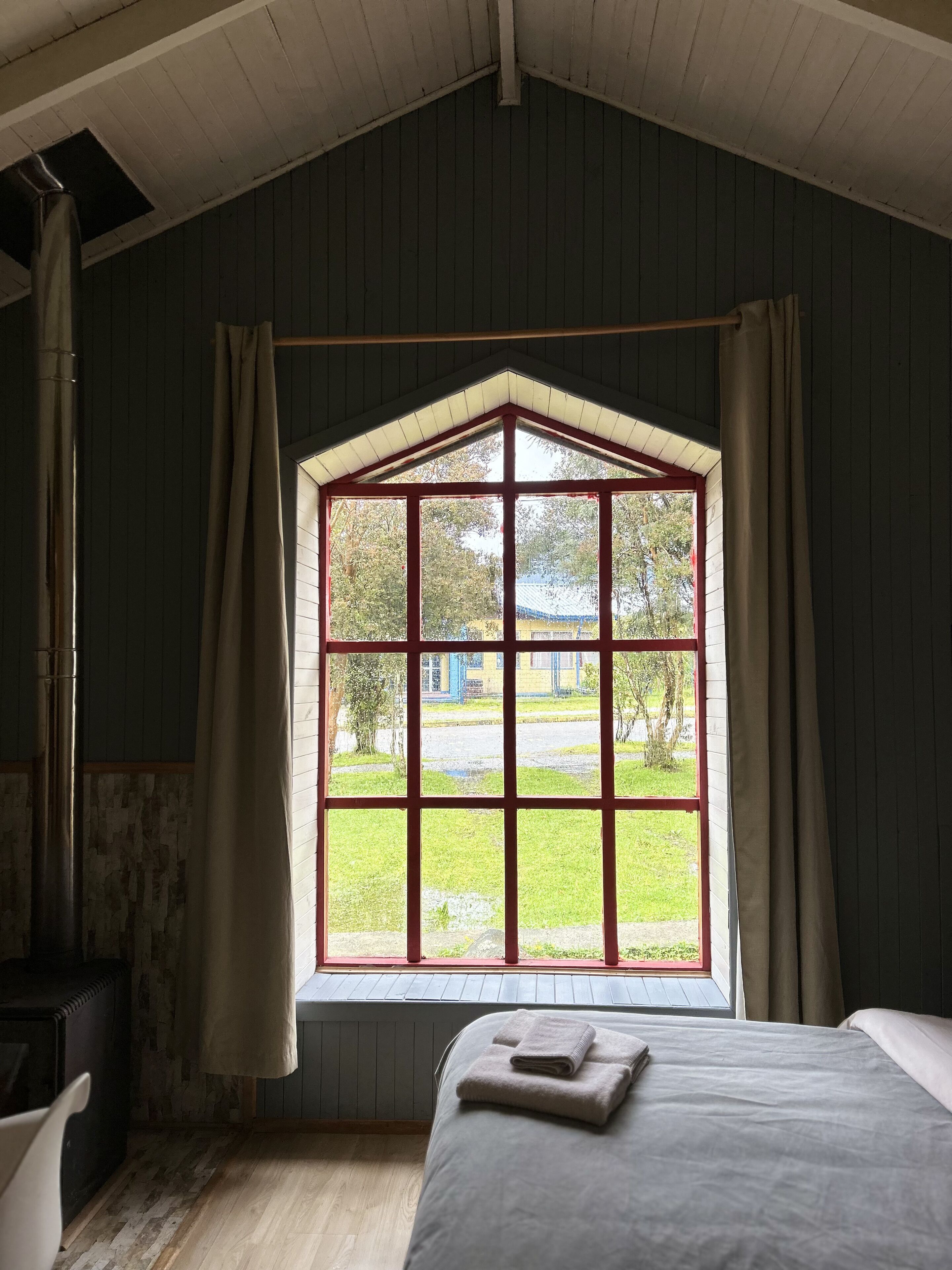 Un dormitorio con una ventana grande, paredes de madera, una cama y una silla.