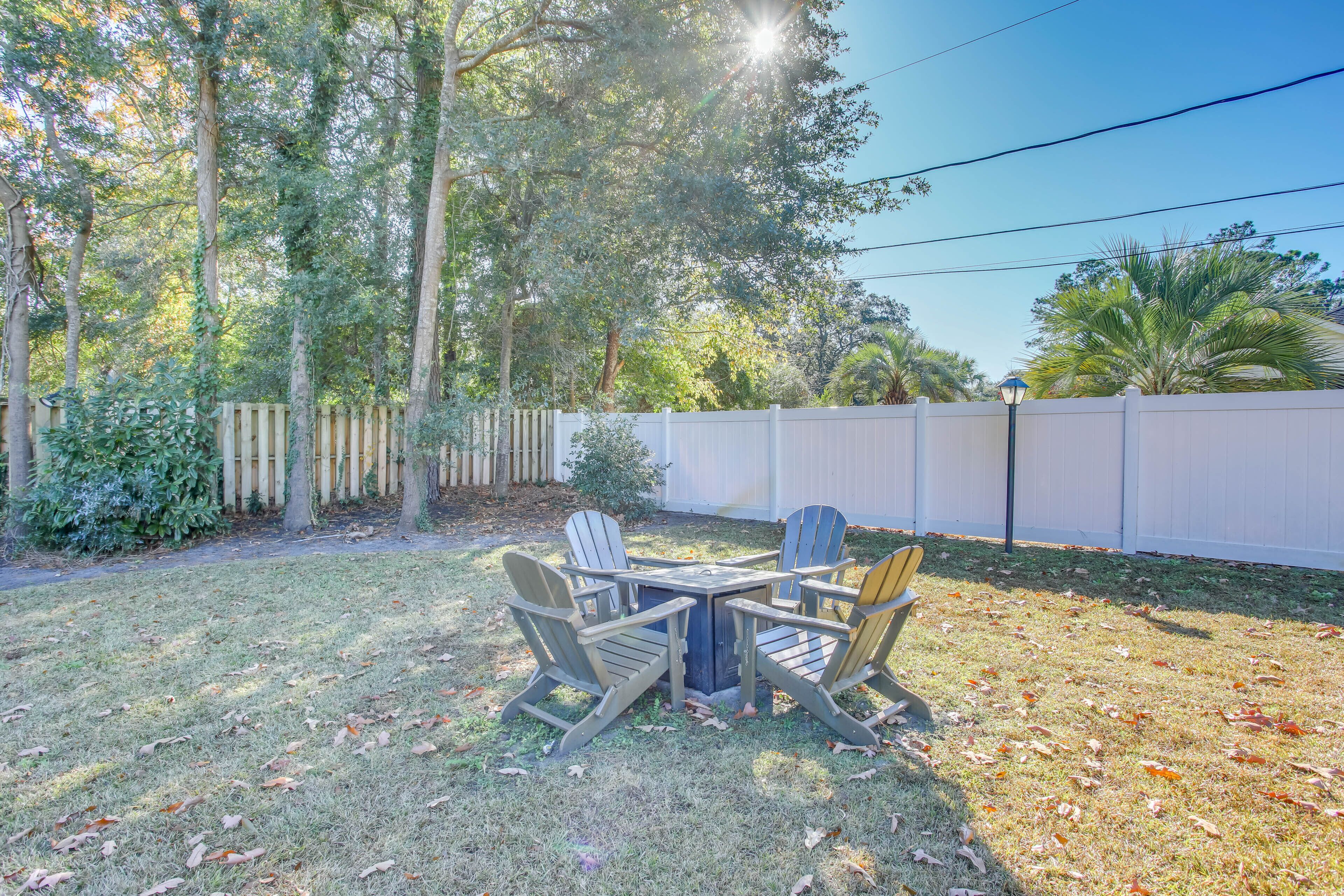 A backyard with a wooden table and four Adirondack chairs, surrounded by a white fence and trees.