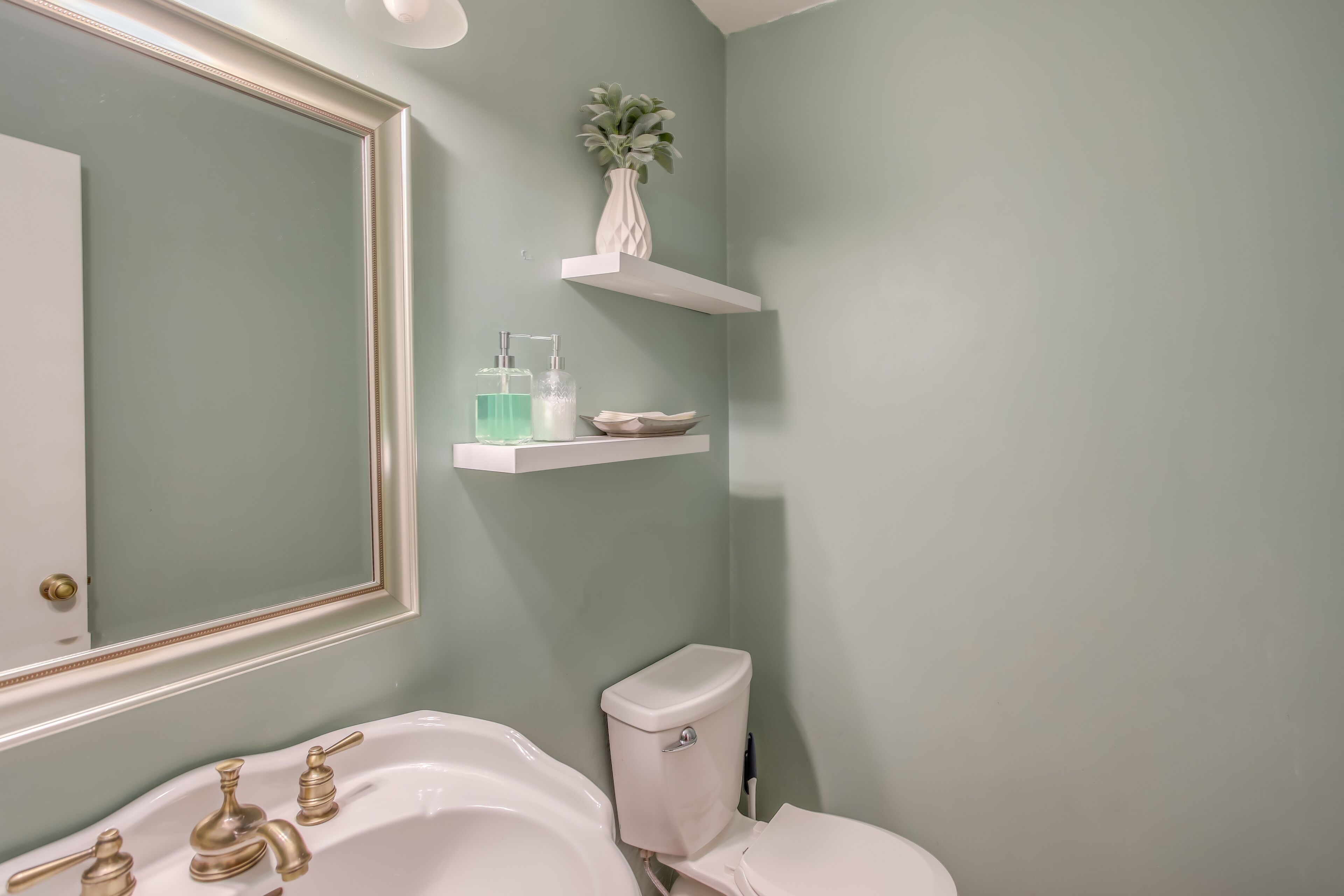 Un bagno con un lavandino bianco, rubinetti dorati, un water e uno specchio. Sopra il water ci sono due mensole bianche: una con un vaso e l'altra con un dispenser di sapone.